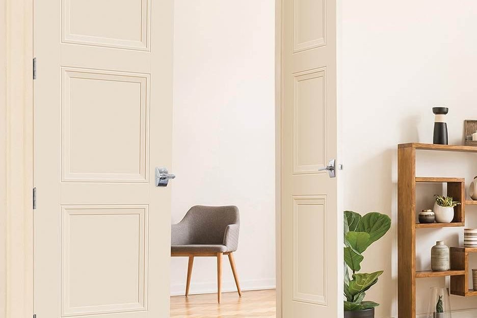 A pair of cream panelled interior doors standing open onto a room with a grey armchair, over herringbone wood flooring