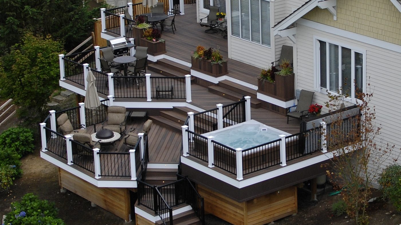 A multi level deck with white railings and steps wrapping the back of a house, with a hot tub on the upper level