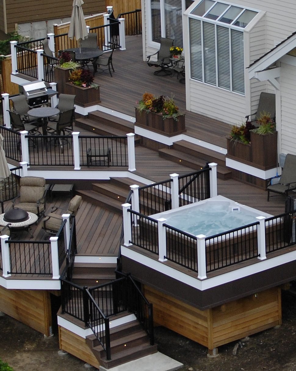 A multi level deck with white railings and steps wrapping the back of a house, with a hot tub on the upper level