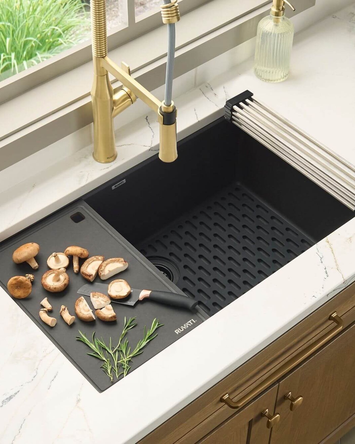 A black composite undermount sink with a brushed gold pull-down faucet, a sliding cutting board and a roll-up drying rack, set in a white marble counter