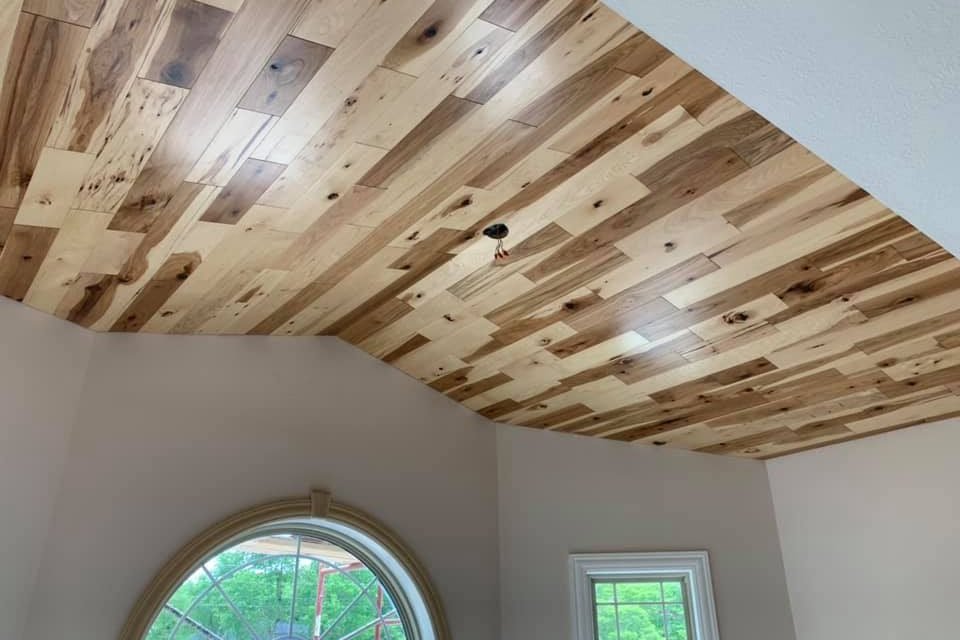 Hickory planks in mixed light and dark tones, laid across a vaulted ceiling that steps down to white walls, above an arched window