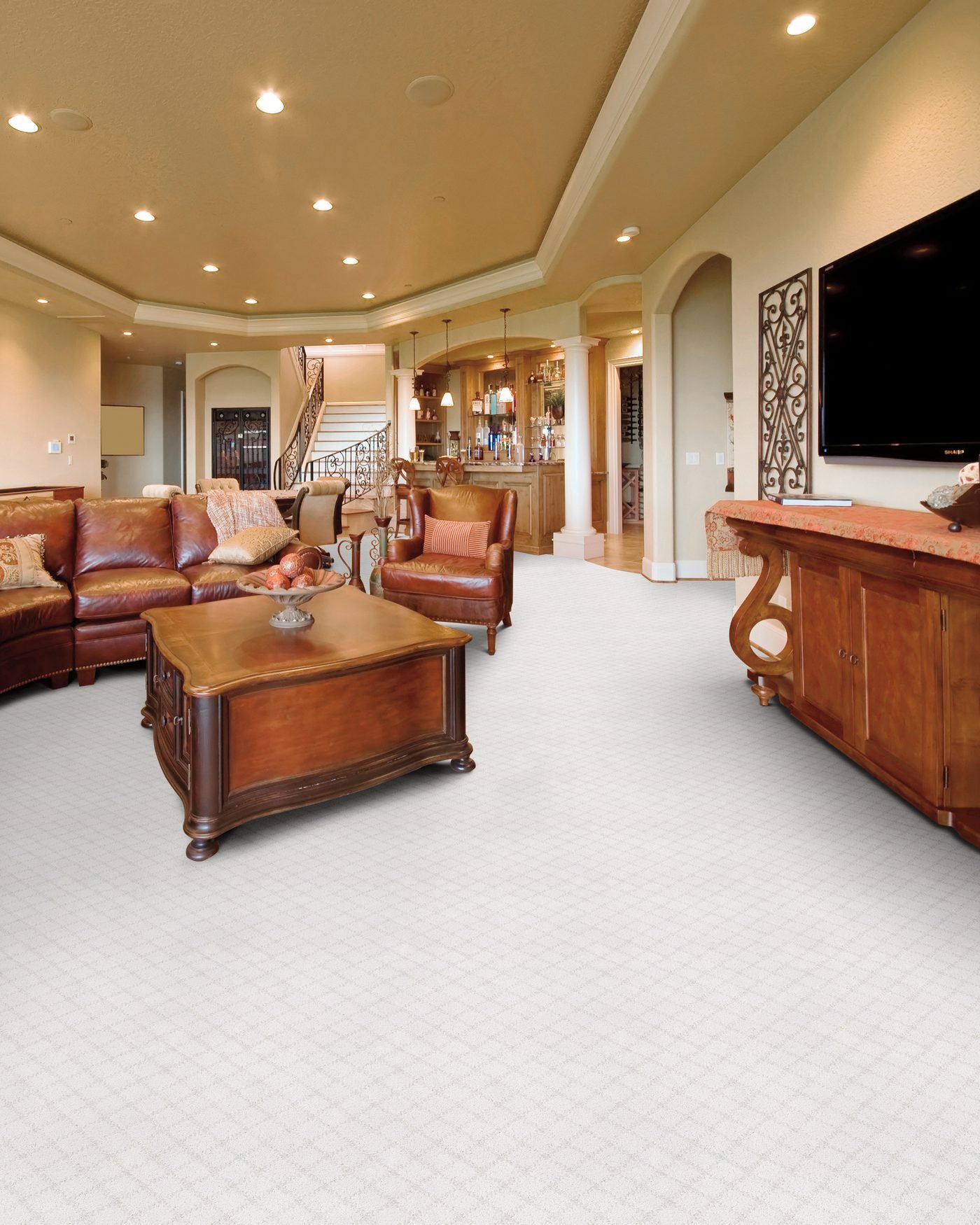 A large finished lower level carpeted wall to wall in a patterned cream loop, furnished with brown leather seating and dark wood, with a bar and a staircase beyond
