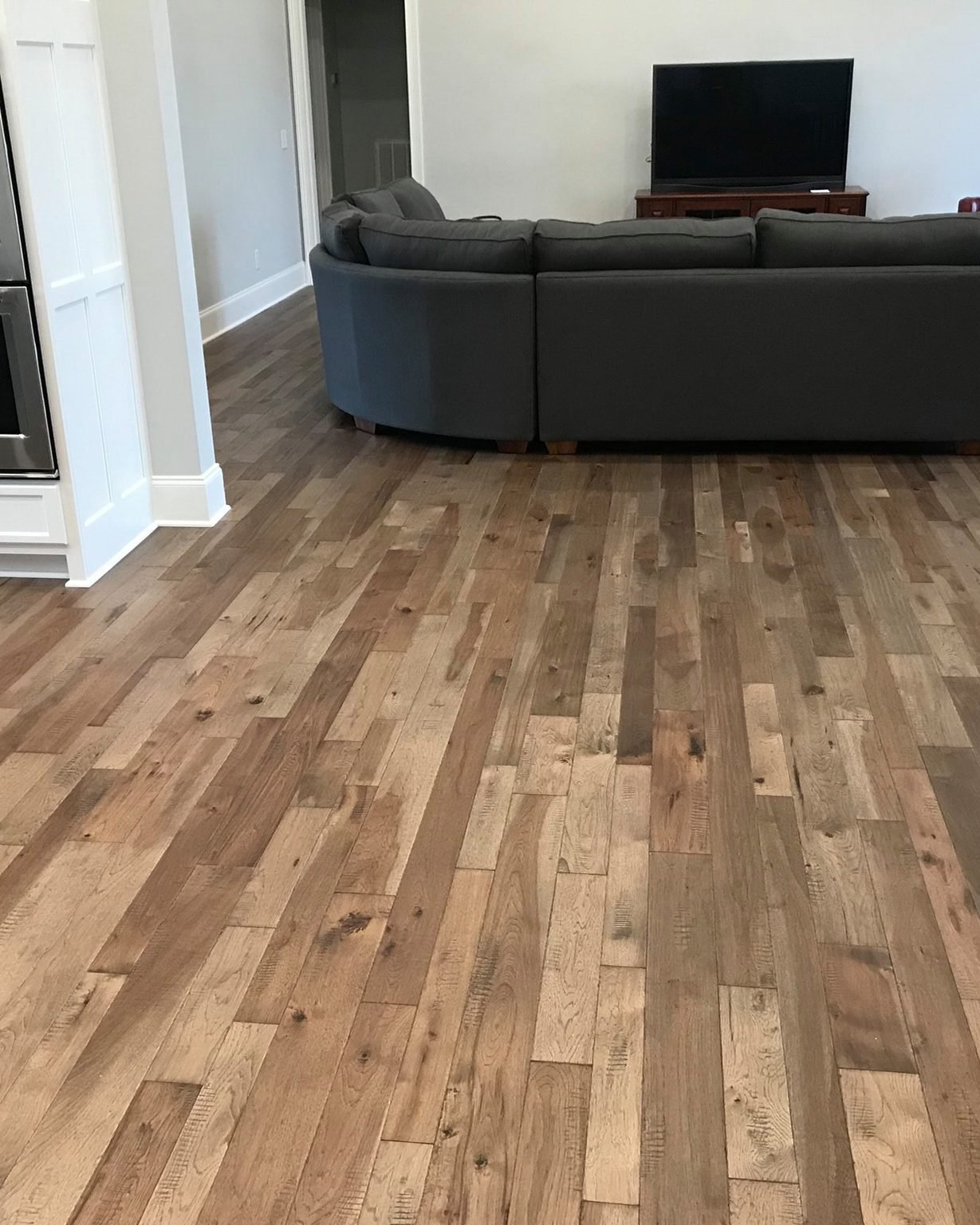 Wide plank hardwood in mixed light and mid browns, the grain and knots clearly visible, running through an open living room with a grey sectional sofa and a television on a dark wood console, past a white kitchen island