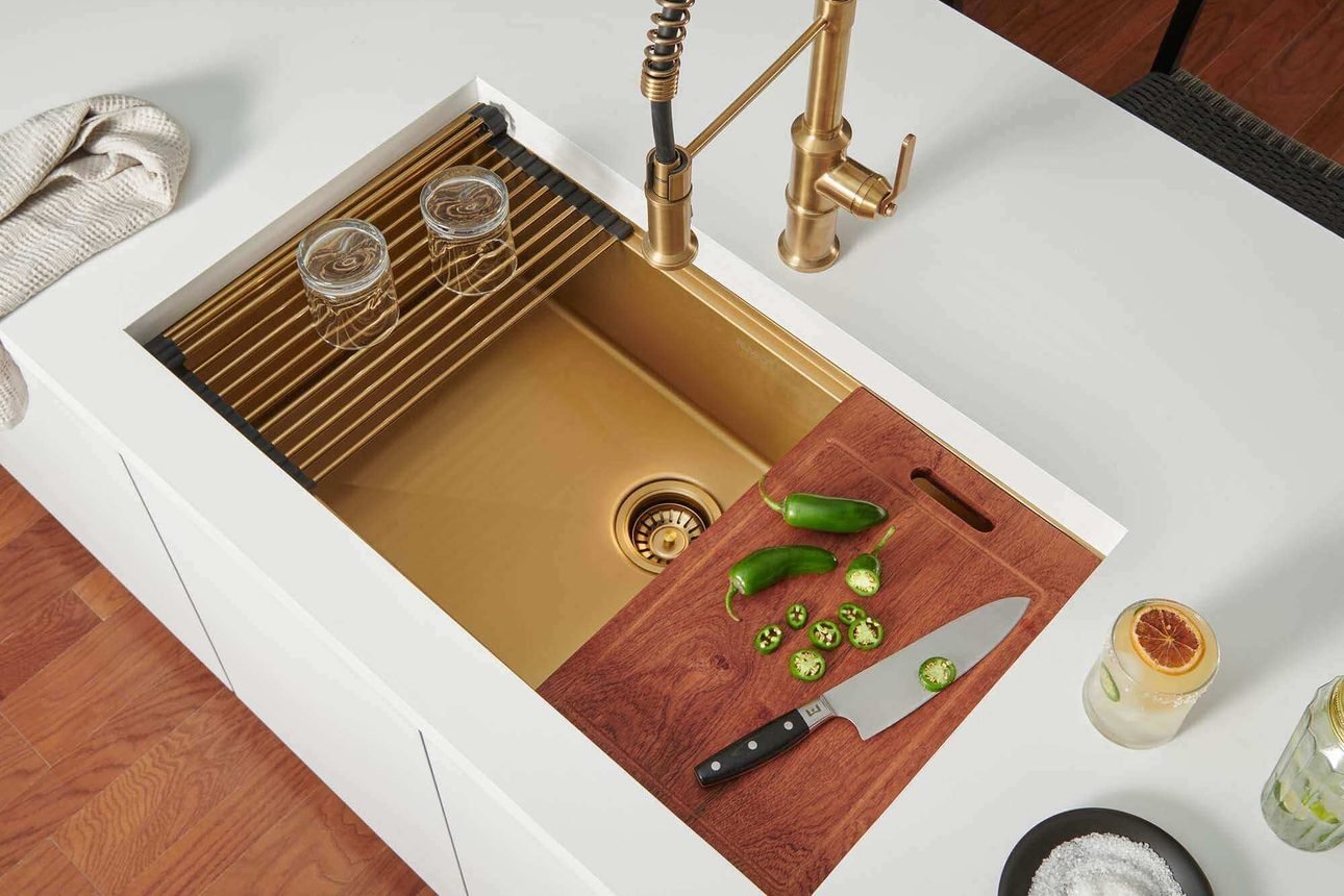 A brushed gold workstation sink set in a white counter, with a matching pull down faucet, a sliding wood cutting board and a roll up drying rack