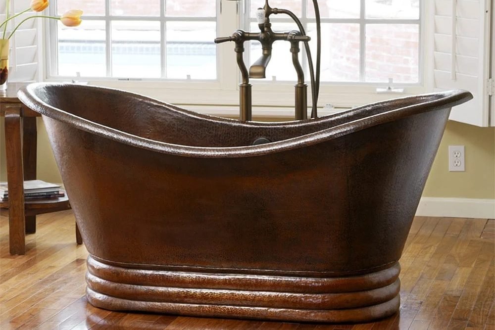A hammered copper double ended freestanding bathtub in an antique finish, on a wood floor beside a brick wall, with a floor mounted telephone tub filler