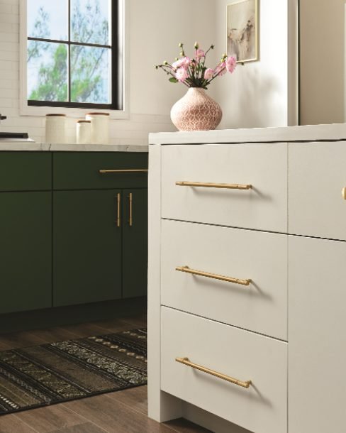 A cream kitchen island with long brushed brass bar pulls and a round knob, in front of a run of deep green cabinets with matching brass hardware