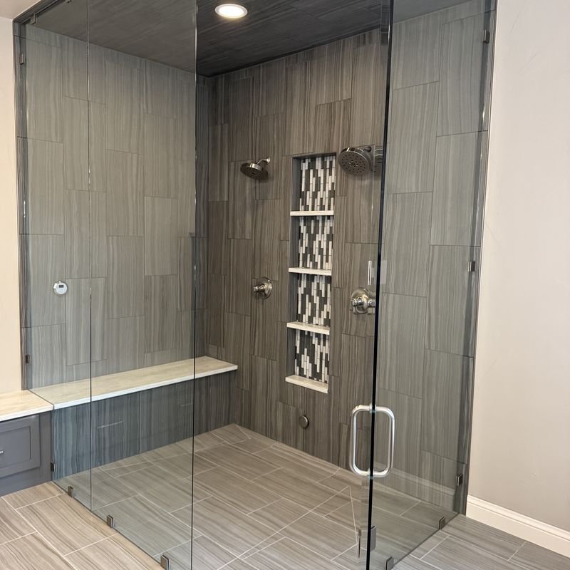 A frameless glass shower tiled floor to ceiling in grey wood-grain porcelain, with a mosaic-lined niche