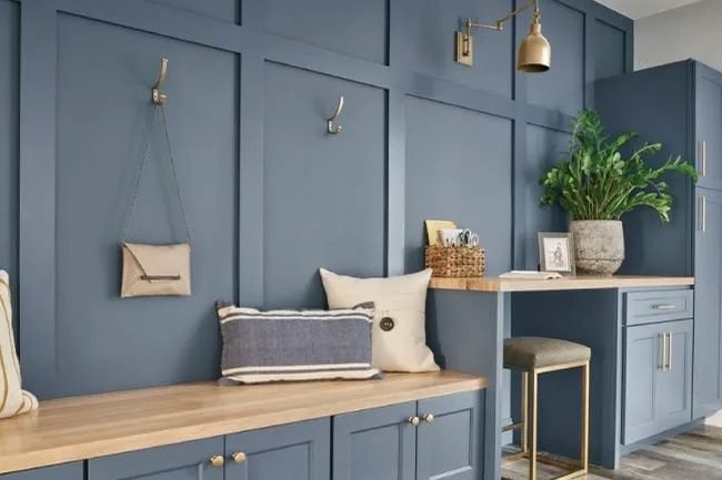 A blue panelled mudroom with a wood bench and coat hooks, beside blue cabinetry with brass pulls under a brass swing arm sconce