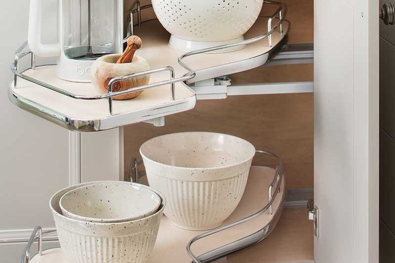 A corner base cabinet standing open on chrome swing out shelves, loaded with bowls, a colander and a kettle