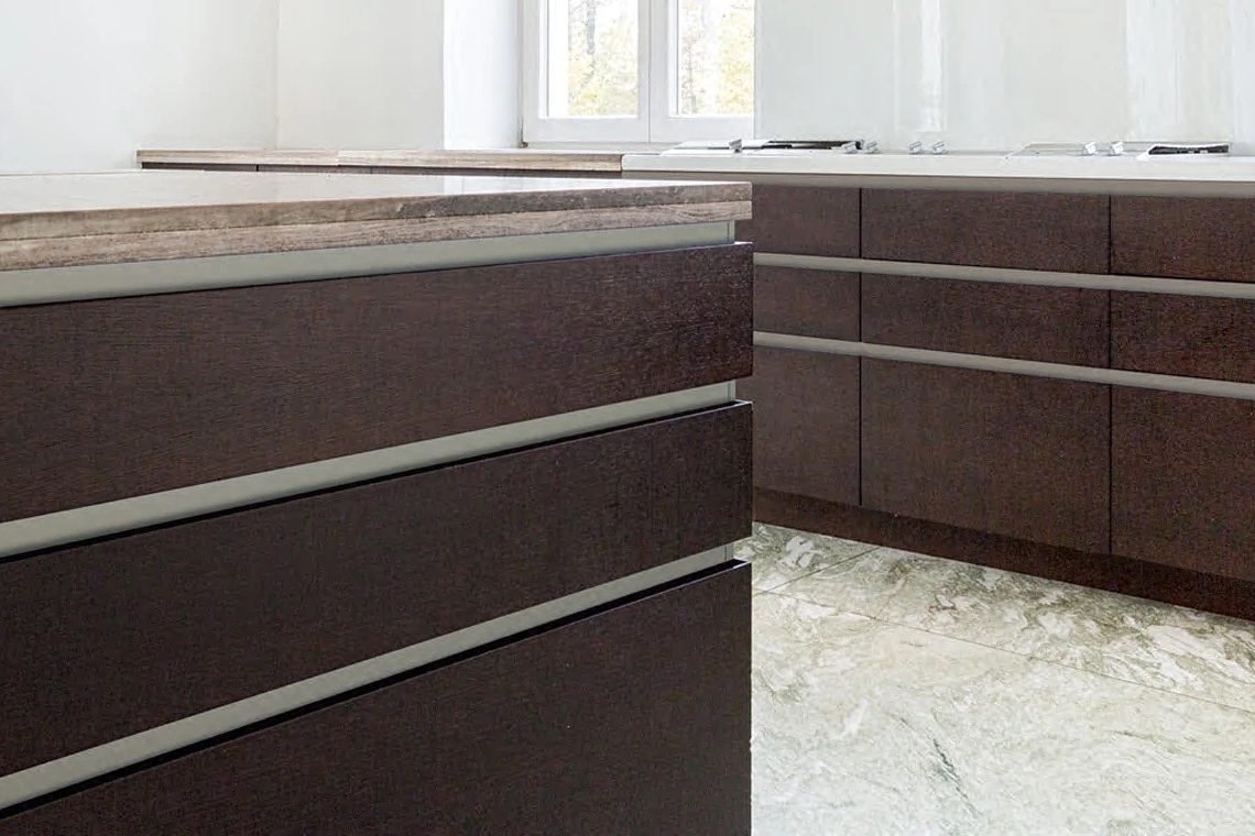 A close view of espresso drawer fronts fitted with long horizontal linear pulls, under a stone counter edge