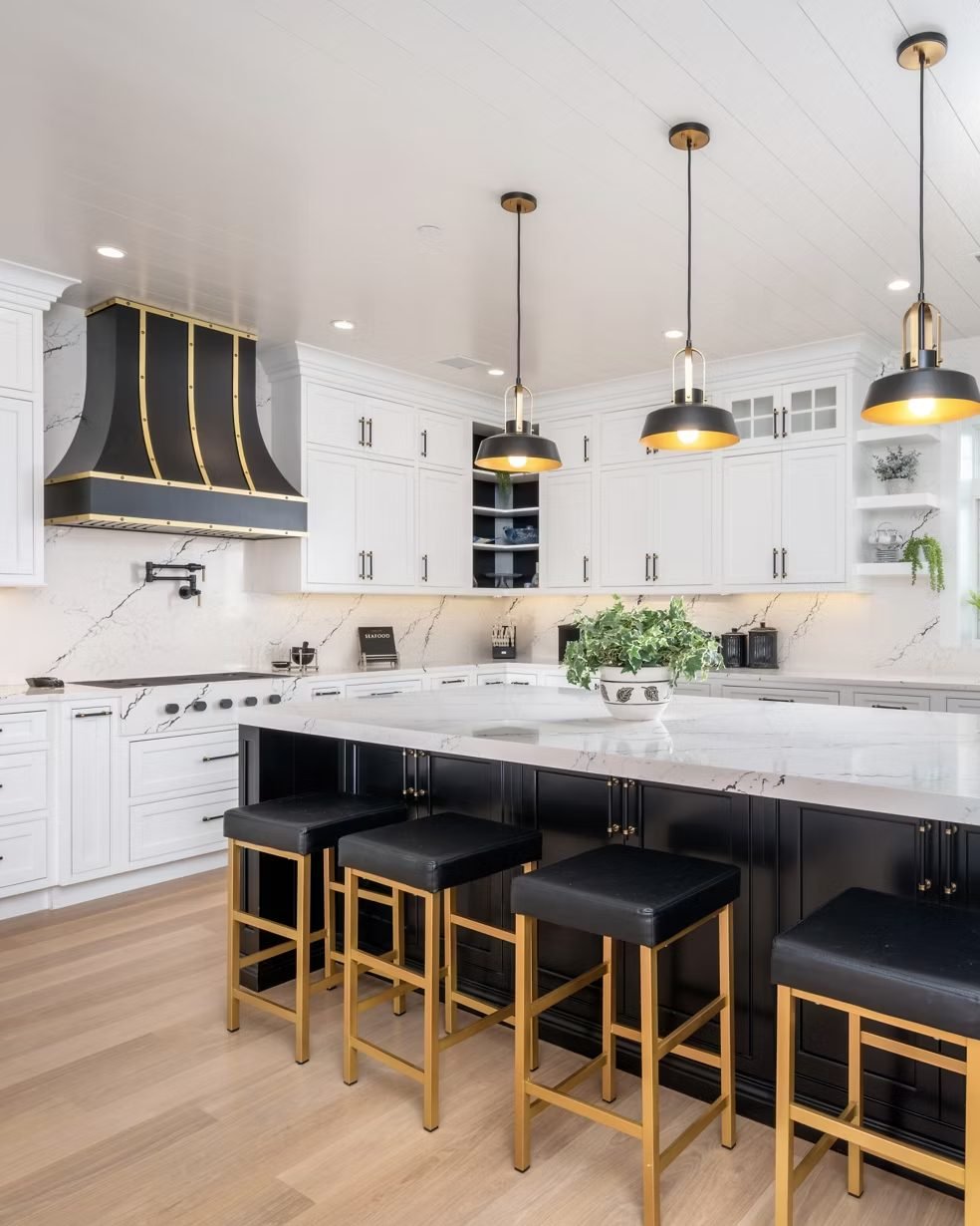 A white kitchen with a dark island, a black and brass hood over the range and stools along the island