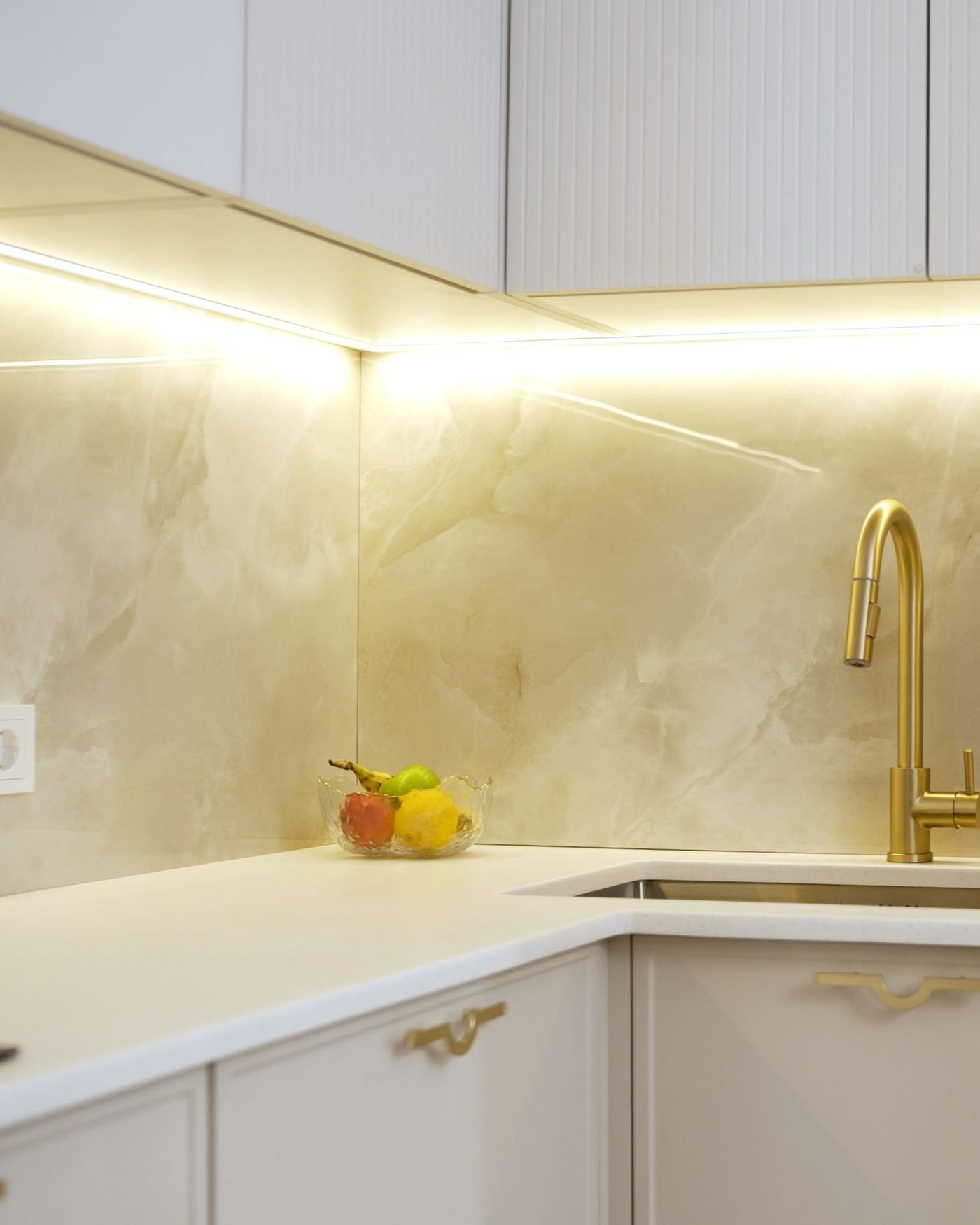 Warm under-cabinet lighting running along two walls of a white kitchen, washing a polished onyx backsplash above a corner counter