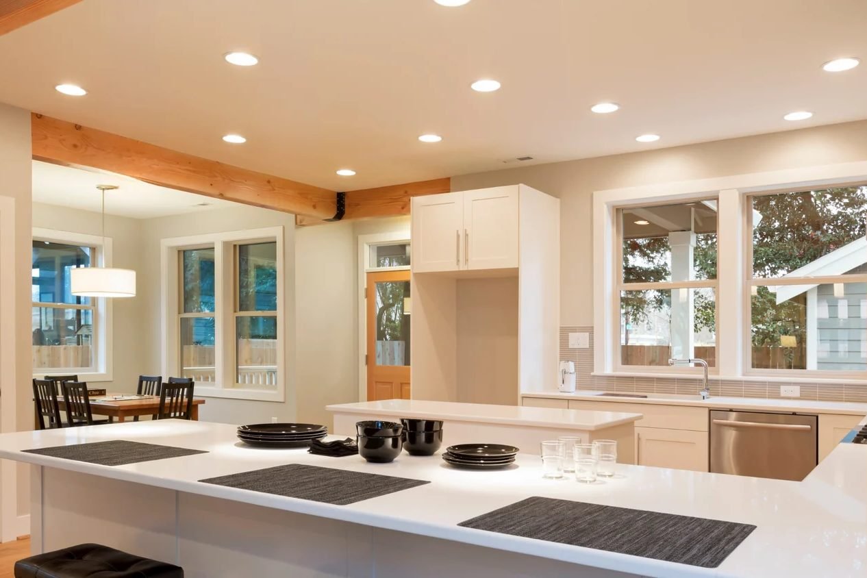 A white kitchen lit by rows of recessed ceiling cans, with a timber beam, a long island and windows onto a dining area