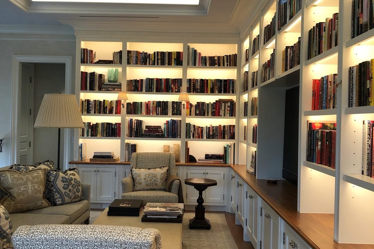 A library wall of white bookshelves lit shelf by shelf with concealed strips, under a tray ceiling washed by cove lighting