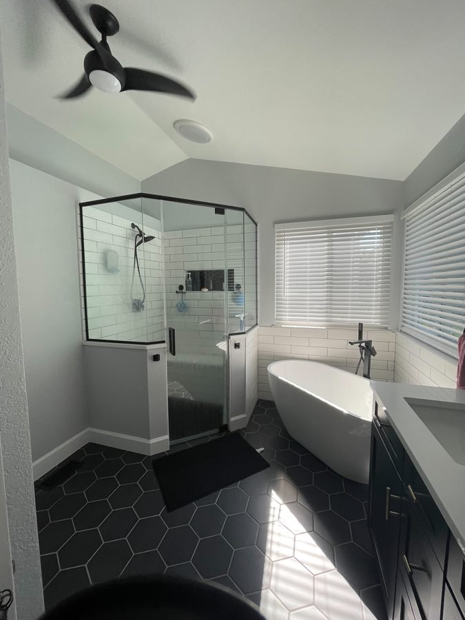A white freestanding tub with a floor-mounted black filler beside a corner glass shower tiled in white subway tile, on a black hexagon tile floor, with a black ceiling fan above and a dark vanity to the right.