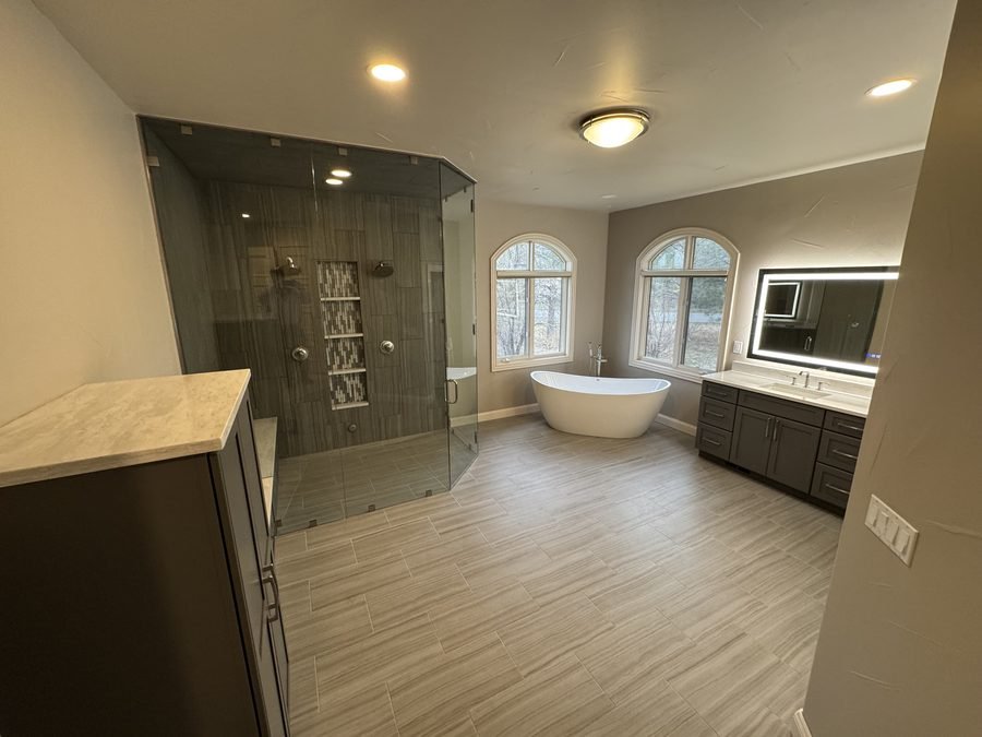 A wide primary bathroom with a white freestanding oval tub set between two arched windows, a large glass shower with a tiled niche on the left, wood-look plank floor and a dark double vanity under a lit mirror.
