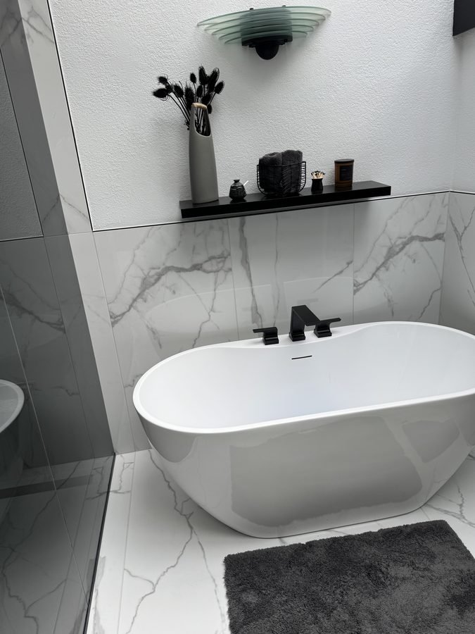 A white freestanding oval tub against a marble-look slab wall, with a matte black deck-mounted filler, a floating black shelf above holding a vase and candles, and a grey bath mat on the floor.