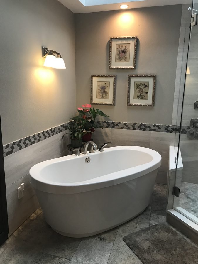 A white freestanding oval tub in a grey tiled alcove with a mosaic border band, a brushed nickel floor-mounted filler, a wall sconce and three framed botanical prints above it, and a glass shower to the right.