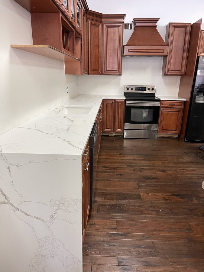 A cherry kitchen with a white stone counter and grey veining, its end panel running down to the floor, with a stainless range under a wood hood on a dark hardwood floor