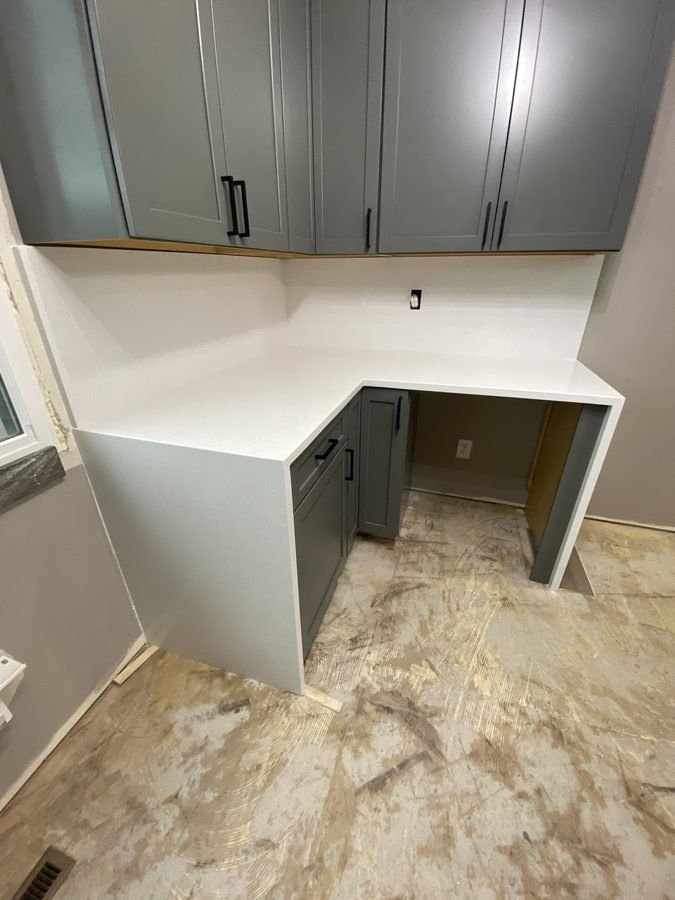 Grey-green cabinets under a white stone counter whose ends run down to the floor on both sides, in a room still unfinished, over a bare subfloor