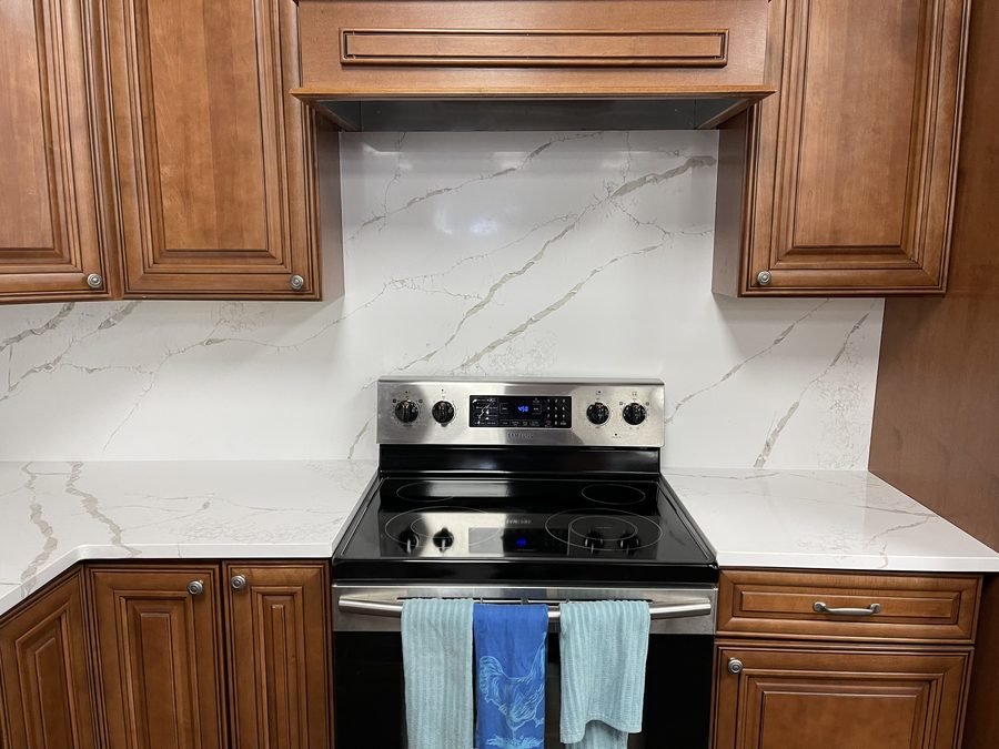 A white stone backsplash with grey veining, running full height behind a stainless range and between cherry cabinets under a wood hood