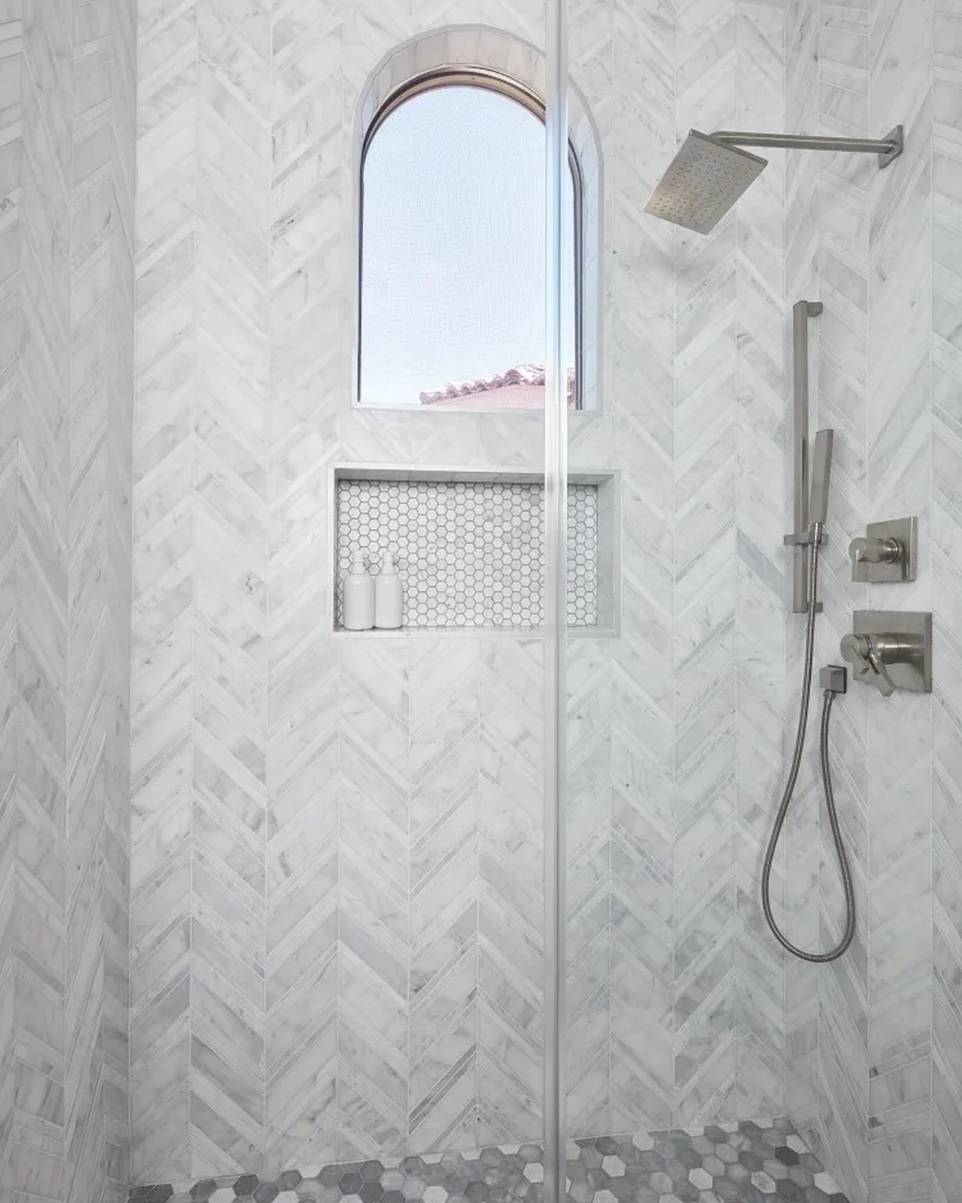 A shower lined in white marble herringbone tile, with a hexagon mosaic niche under an arched window and a grey and white hexagon mosaic floor