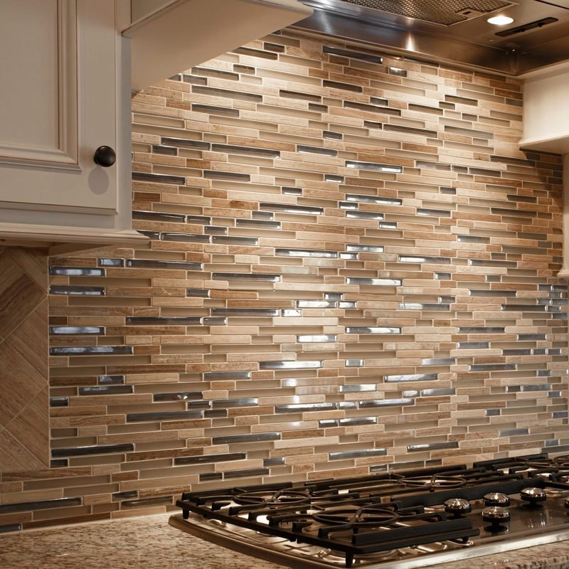 A linear mosaic backsplash in cream and tan stone with glossy glass and metallic strips, running the wall behind a stainless gas cooktop under a chimney hood, above a speckled granite counter