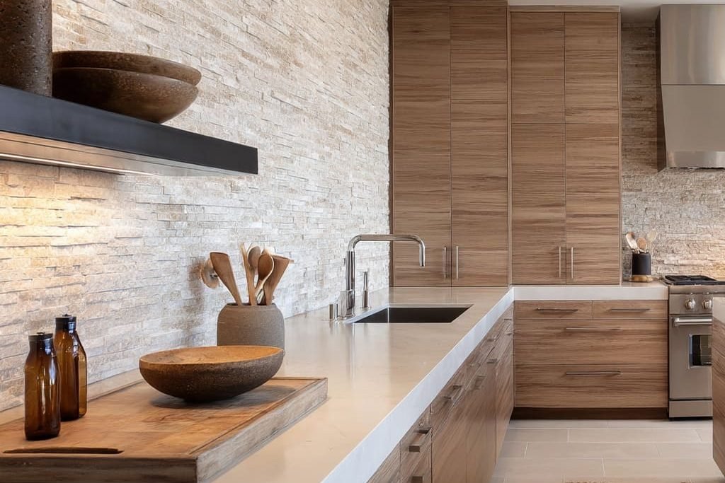 A split face stone backsplash in cream and grey running the full wall of a kitchen in wood cabinetry, behind a white counter and a butcher block ledge