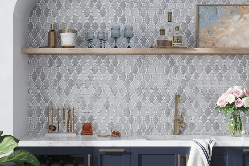 A grey lantern shaped mosaic filling an arched alcove above a marble counter, behind navy cabinetry and a floating wood shelf of glassware