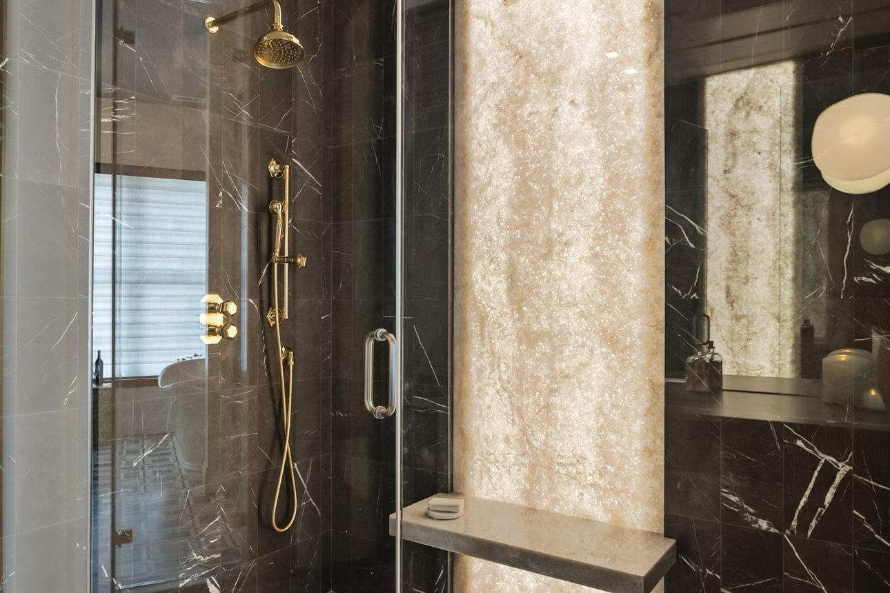 A dark marble shower with brass fittings and a backlit honey onyx panel above a stone bench, over a mosaic floor