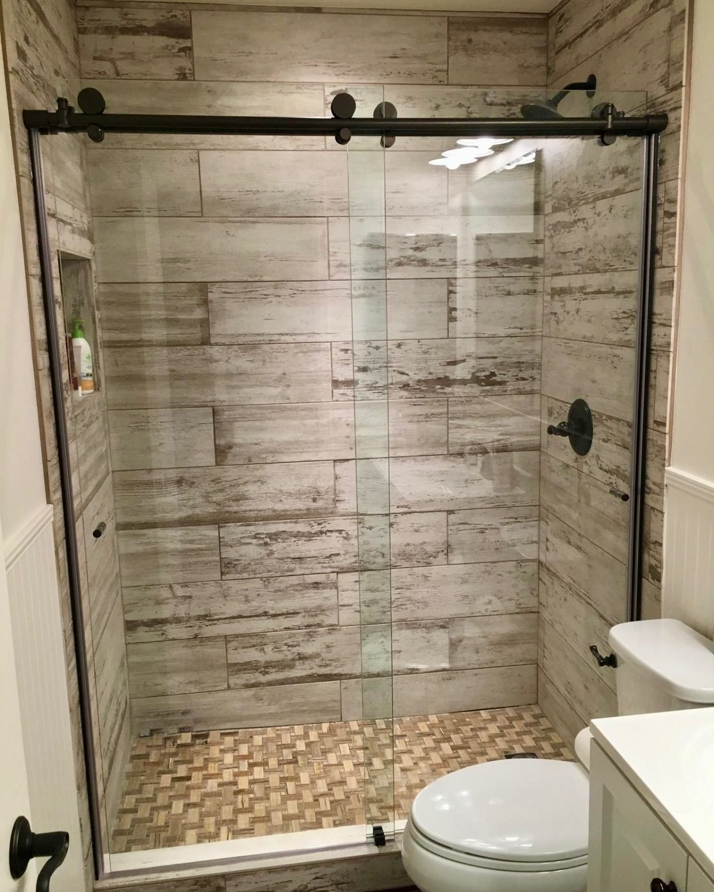 A walk in shower lined in weathered wood-look plank tile, behind a black barn-style sliding glass door, over a woven basketweave tile floor