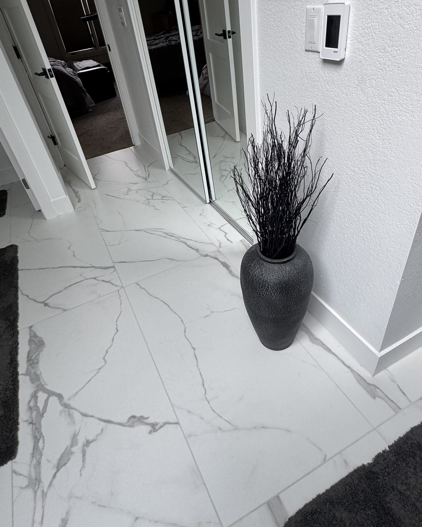 A bathroom floor in large format white marble-look porcelain running out through an open doorway, the veining carrying across the joints, with a tall black textured vase standing against the tiled wall