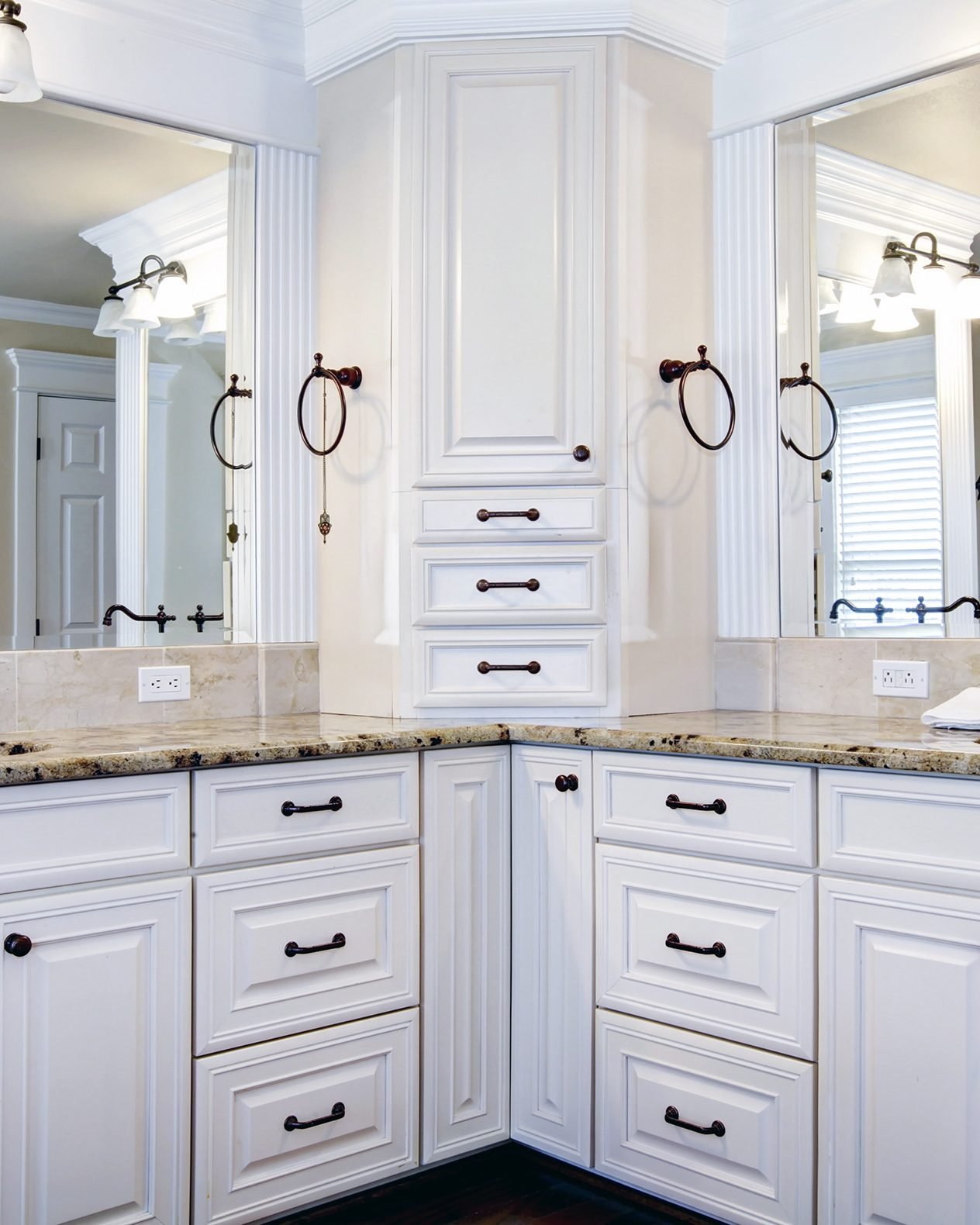 A white double vanity run into a corner, with a tall storage tower between two framed mirrors, a speckled granite top, oil rubbed bronze faucets and towel rings, under two glass shade light bars