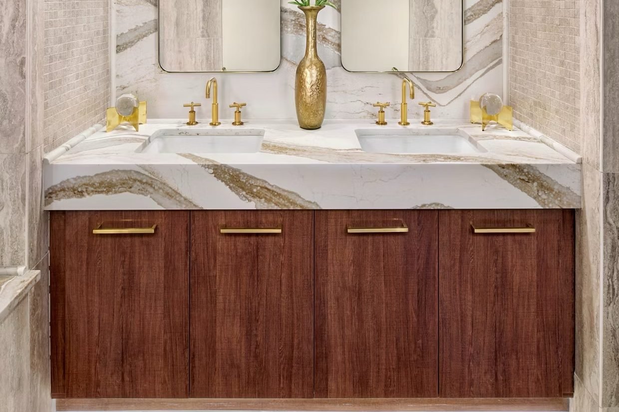 A floating walnut double vanity with brass cross-handle faucets, under two rounded mirrors on a veined stone wall, lit from below