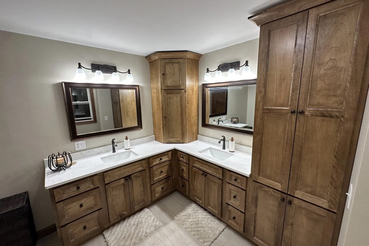 An L-shaped stained maple double vanity with white quartz tops, framed mirrors, a corner storage tower and a full-height linen cabinet