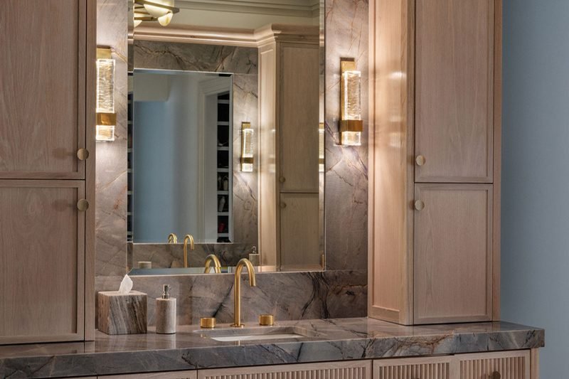 A pale oak vanity with fluted drawer fronts between two tall storage towers, under a mirror flanked by lit brass sconces, with a grey veined stone top and full height surround
