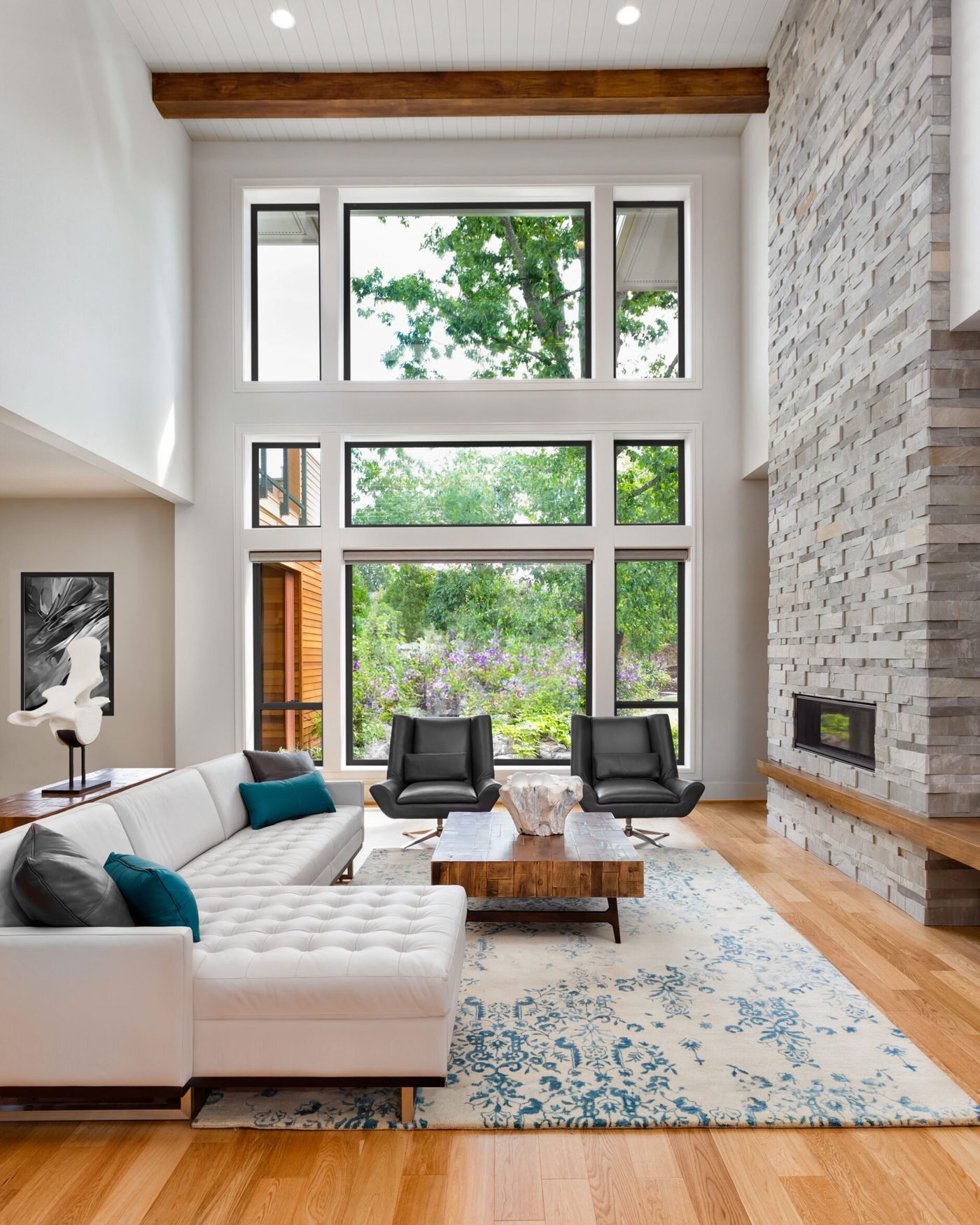 A double height living room walled in black framed windows looking onto trees, beside a stacked stone fireplace, under a timber ceiling beam