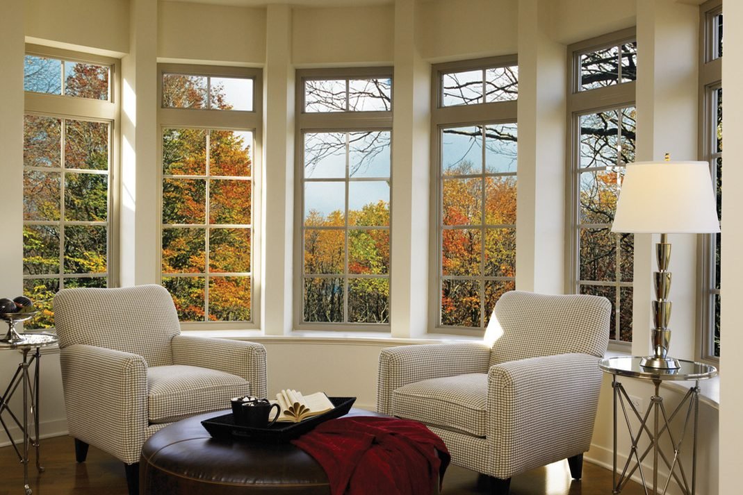 A curved bay of white grilled windows around a sitting area with two cream armchairs and a lamp, looking onto autumn trees