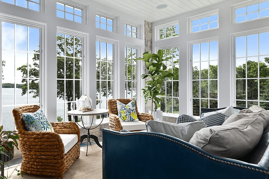 A sunroom glazed on three walls under a white plank ceiling, furnished with a navy sofa, woven armchairs and a potted palm