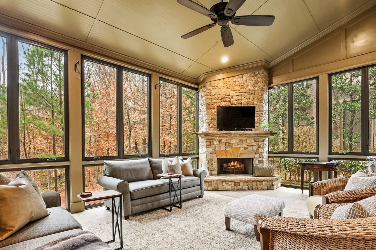 A screened porch walled in black framed windows onto bare winter woods, with a stone corner fireplace, a vaulted board ceiling and a ceiling fan